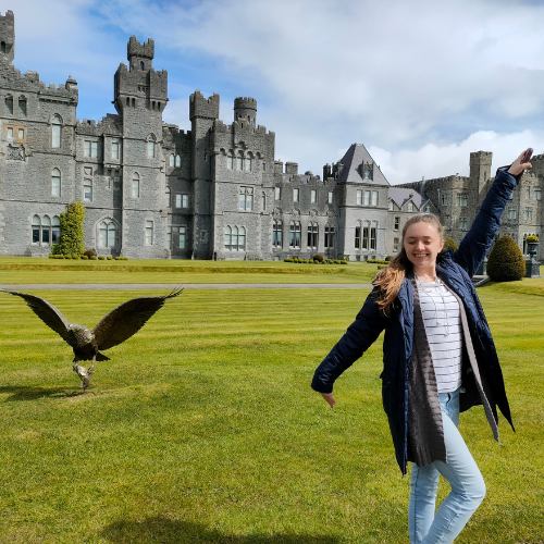 Young woman stands in front of castle with a large bird of prey flying low in the background.